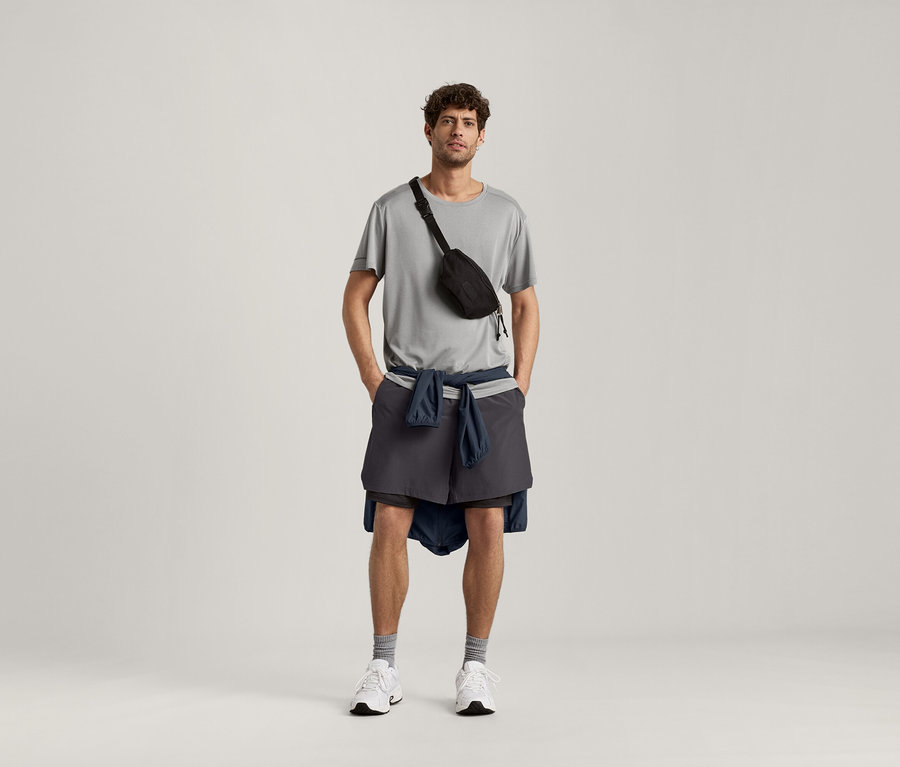 Muž má na sebe svetlosivé melírované športové tričko, čiernu tašku na bedrá, šortky a bundu uviazanú okolo pása.