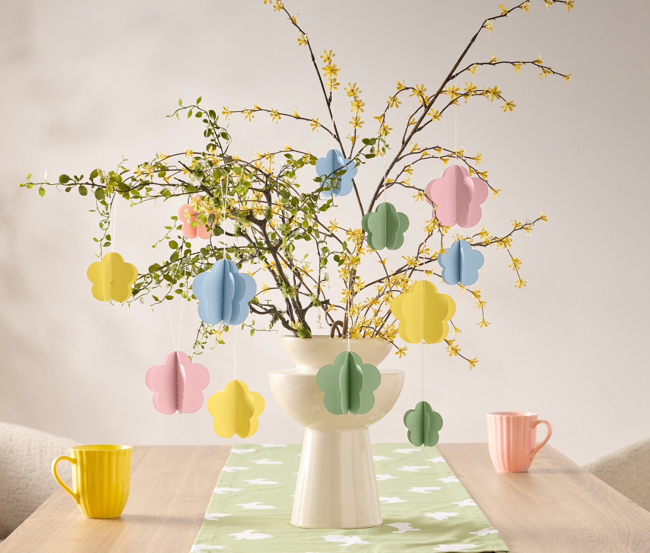 Kvetinové ozdoby visia na vetve vo váze na stole, vedľa žltého a ružového hrnčeka.