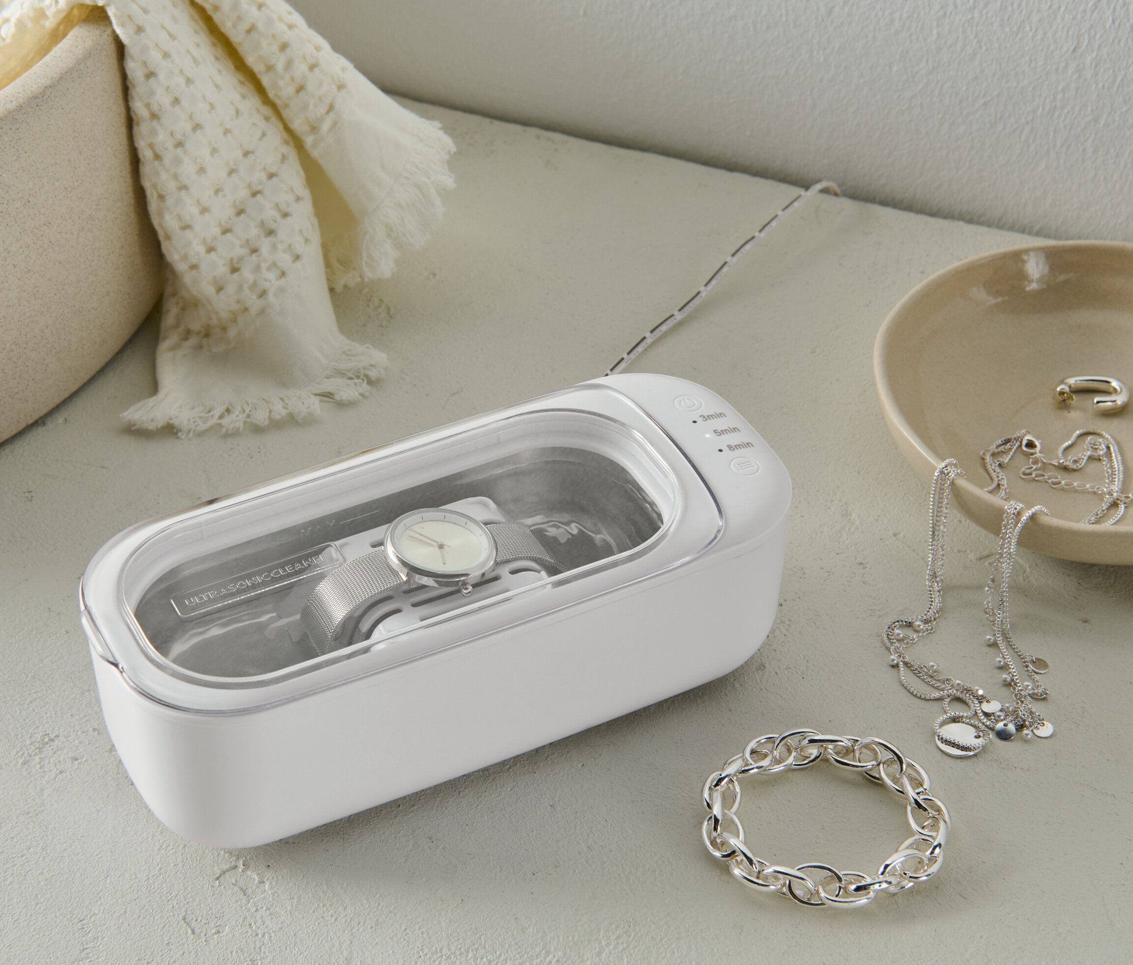 Hodinky sa čistia v bielom ultrazvukovom čističi. Vedľa sú šperky.