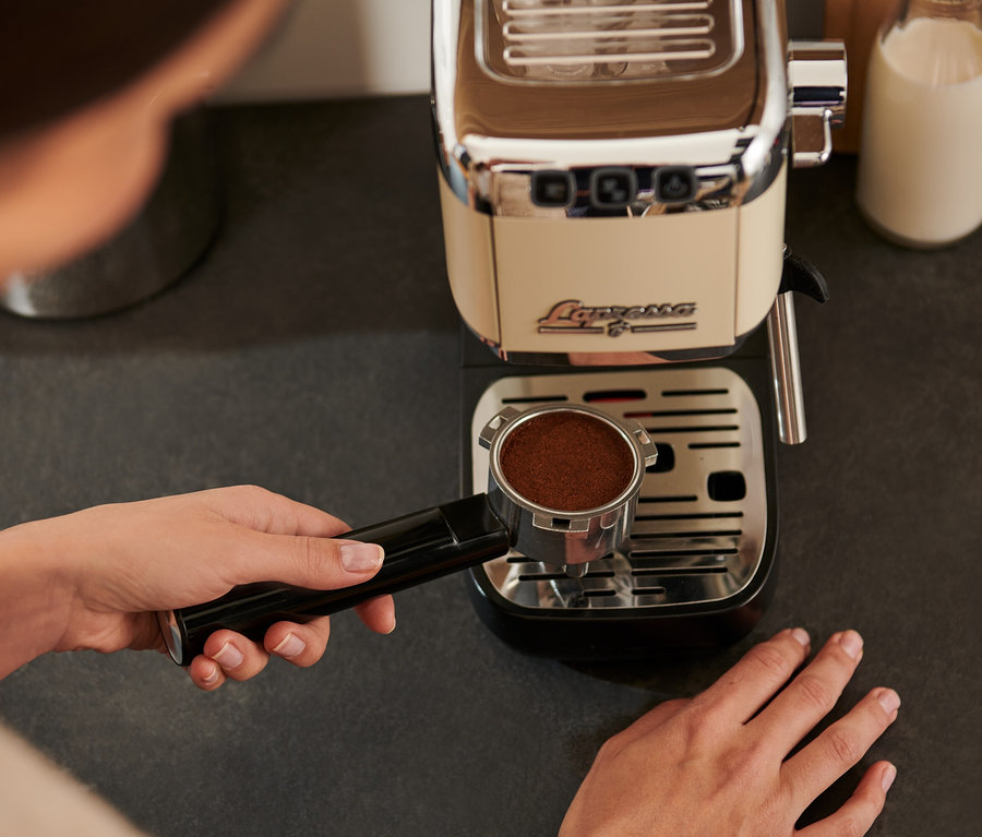 Žena drží páku kávovaru »Lapressa« krémovej farby s namletou kávou nad odkvapkávacou mriežkou.