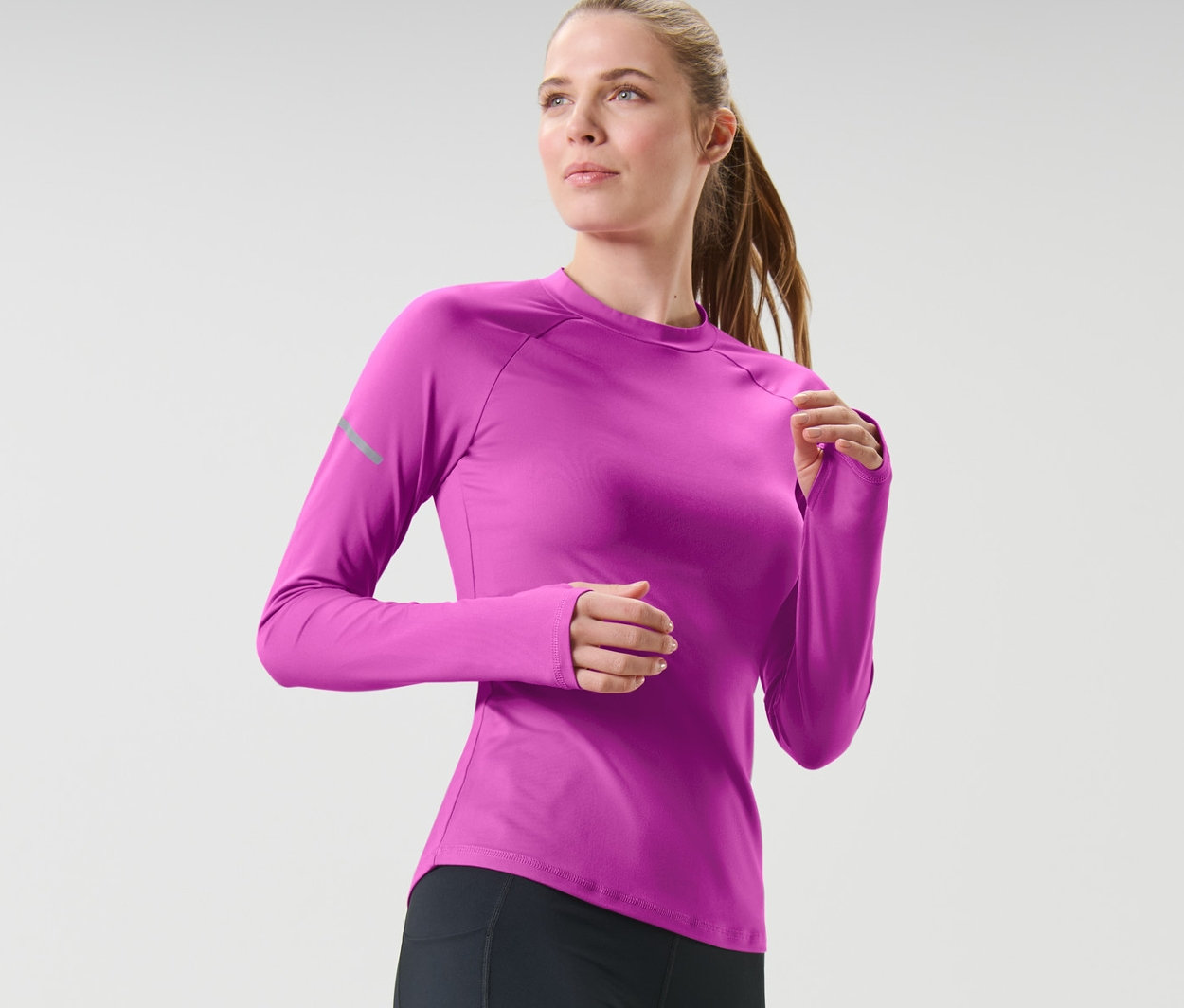 Žena v čiernych legínach a neónovovínovom funkčnom tričku s dlhými rukávmi beží.