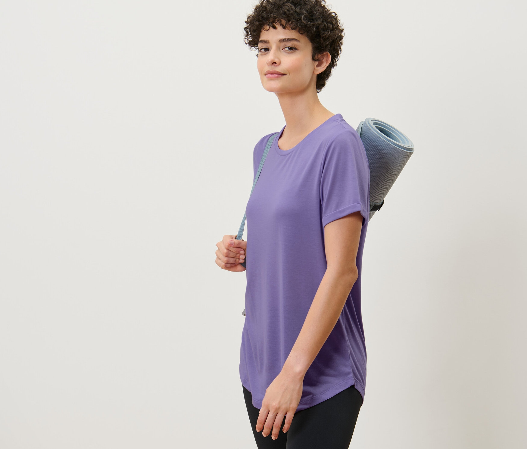 Žena má na sebe fialové dlhé tričko a nesie podložku na cvičenie.