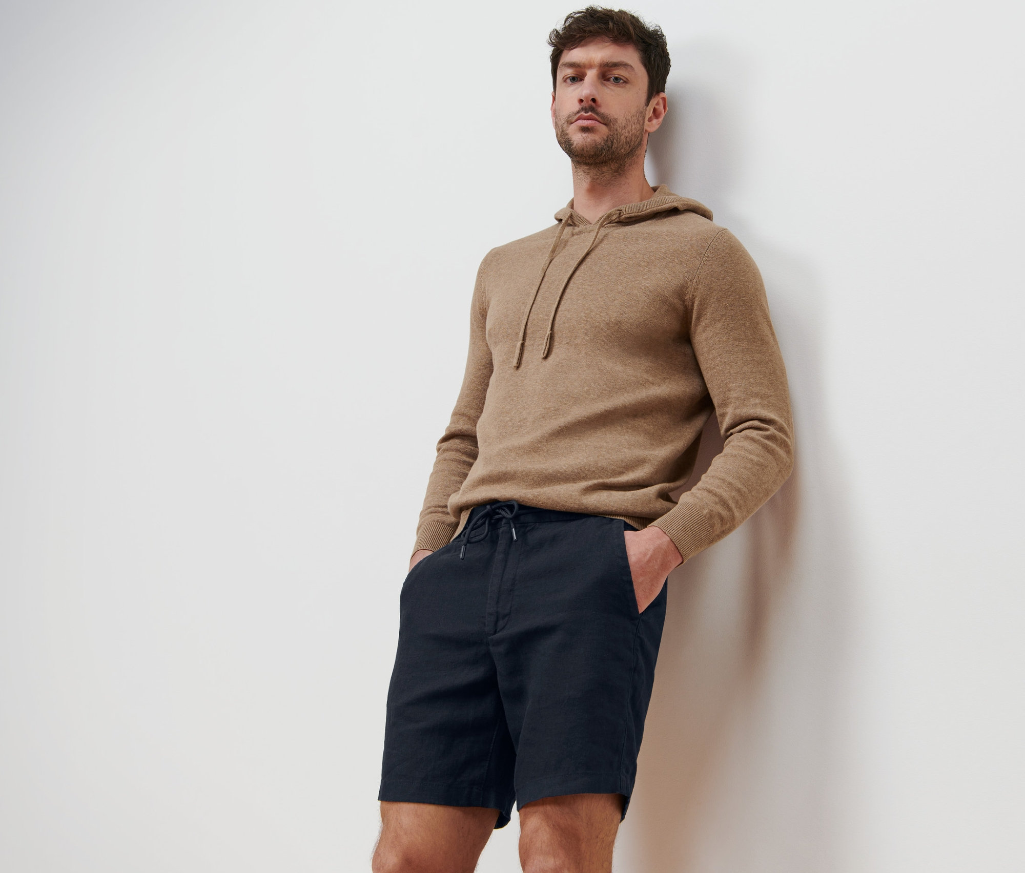 Muž opretý o stenu má na sebe ľanové šortky a mikinu s kapucňou.