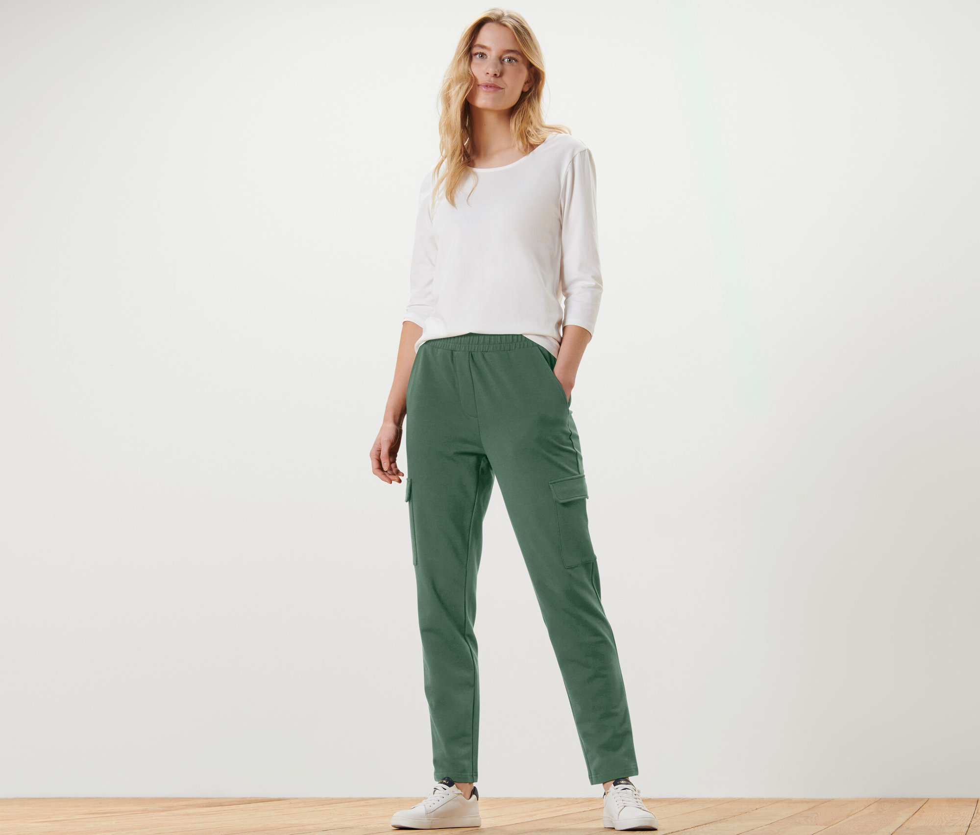 Žena pózuje v zelených teplákových nohaviciach s cargo vreckami a bielom tričku.
