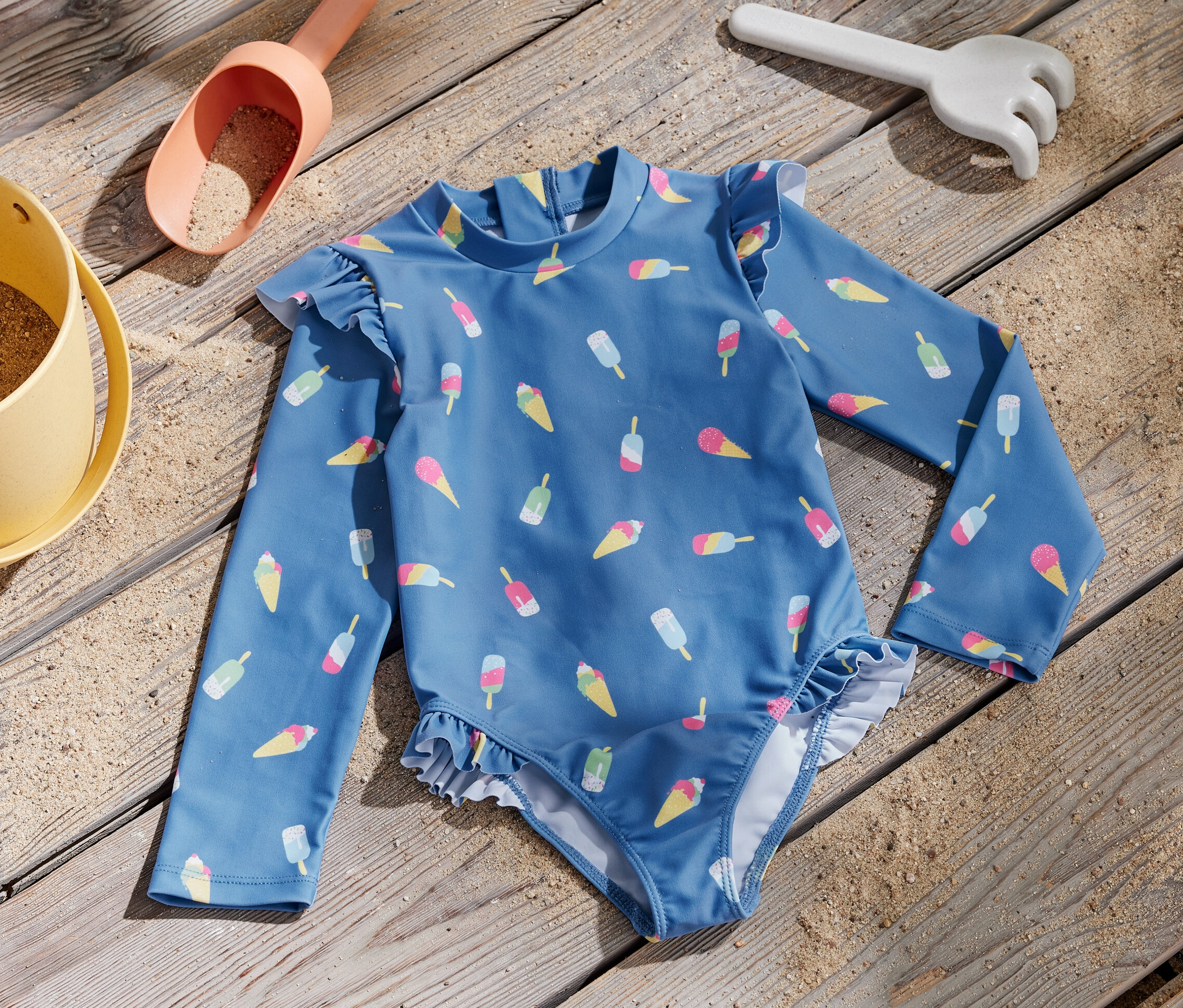 Modré detské plavky s potlačou zmrzliny a UV faktorom 80 ležia na drevenom povrchu vedľa hračky na piesok.