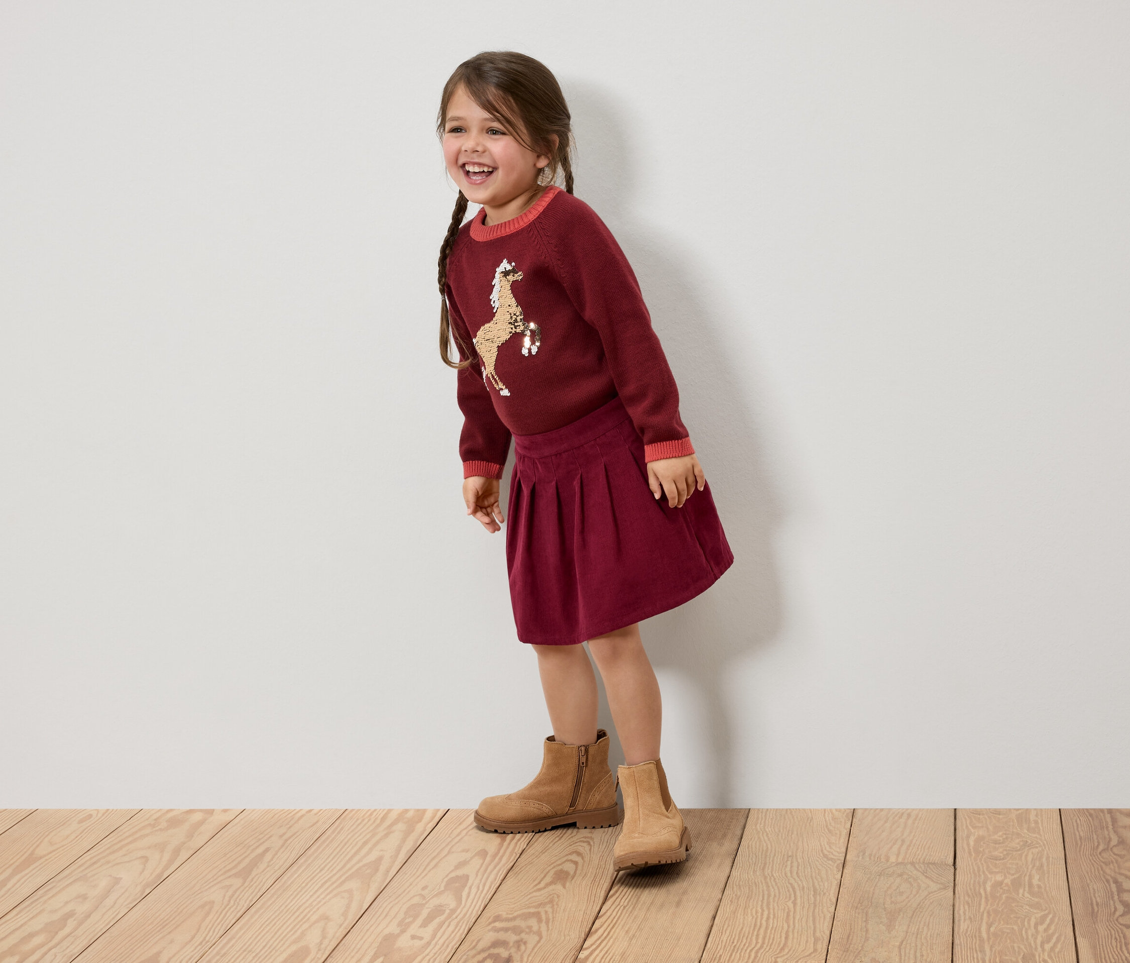 Usmievajúce sa dievčatko s vrkočmi má na sebe bordový sveter s koňom, sukňu a hnedé topánky.