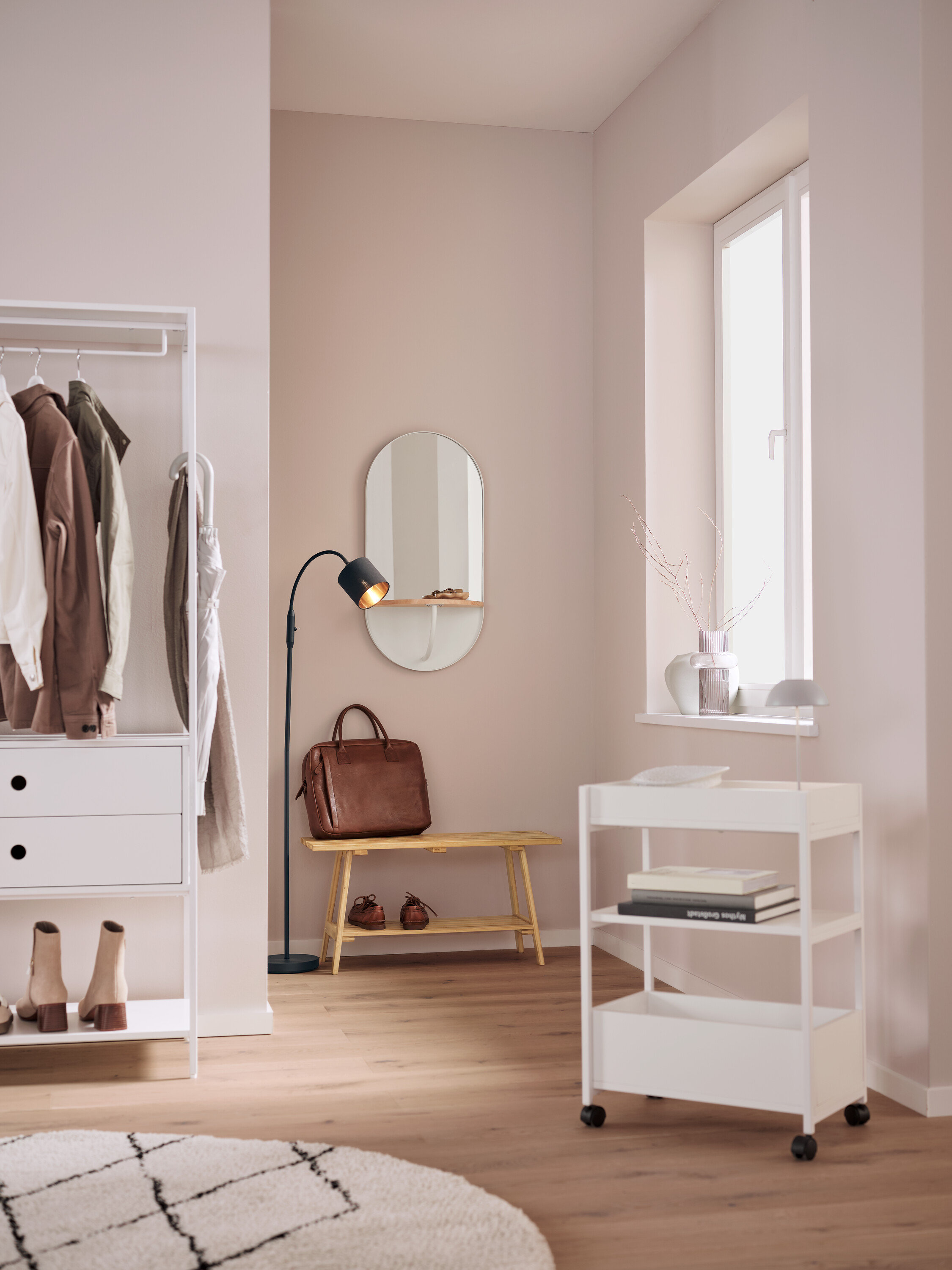 V interiéri sa nachádza biely regál s oblečením, oválne zrkadlo, hnedá kožená taška na lavici, čierna lampa a biely pojazdný vozík.