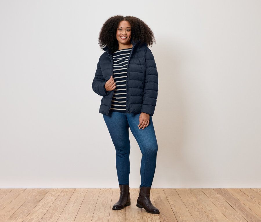 Žena s kučeravými vlasmi stojí a má na sebe modrú prešívanú bundu, pruhované tričko, džínsy a topánky.