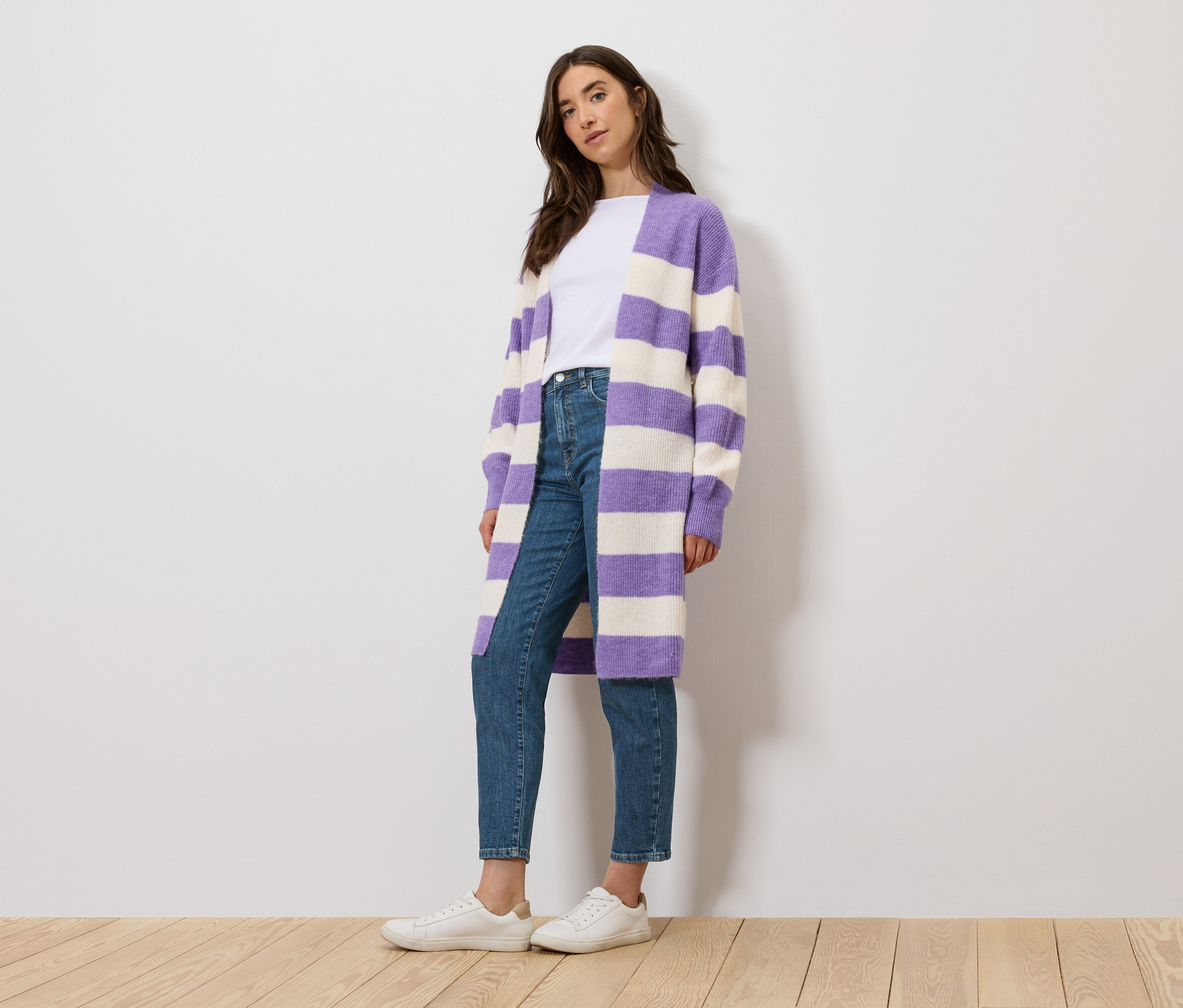 Žena stojí a má na sebe biele tričko, džínsy a fialový a biely pruhovaný kardigán.