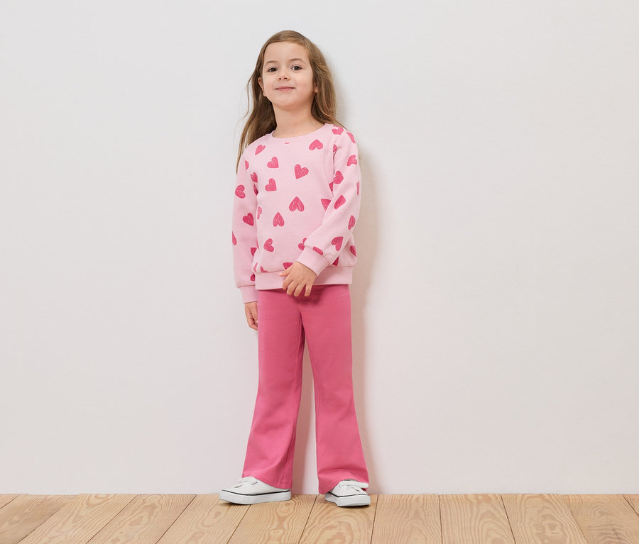 Dievča stojí pred bielou stenou v ružovom svetri so srdiečkami a ružových nohaviciach.