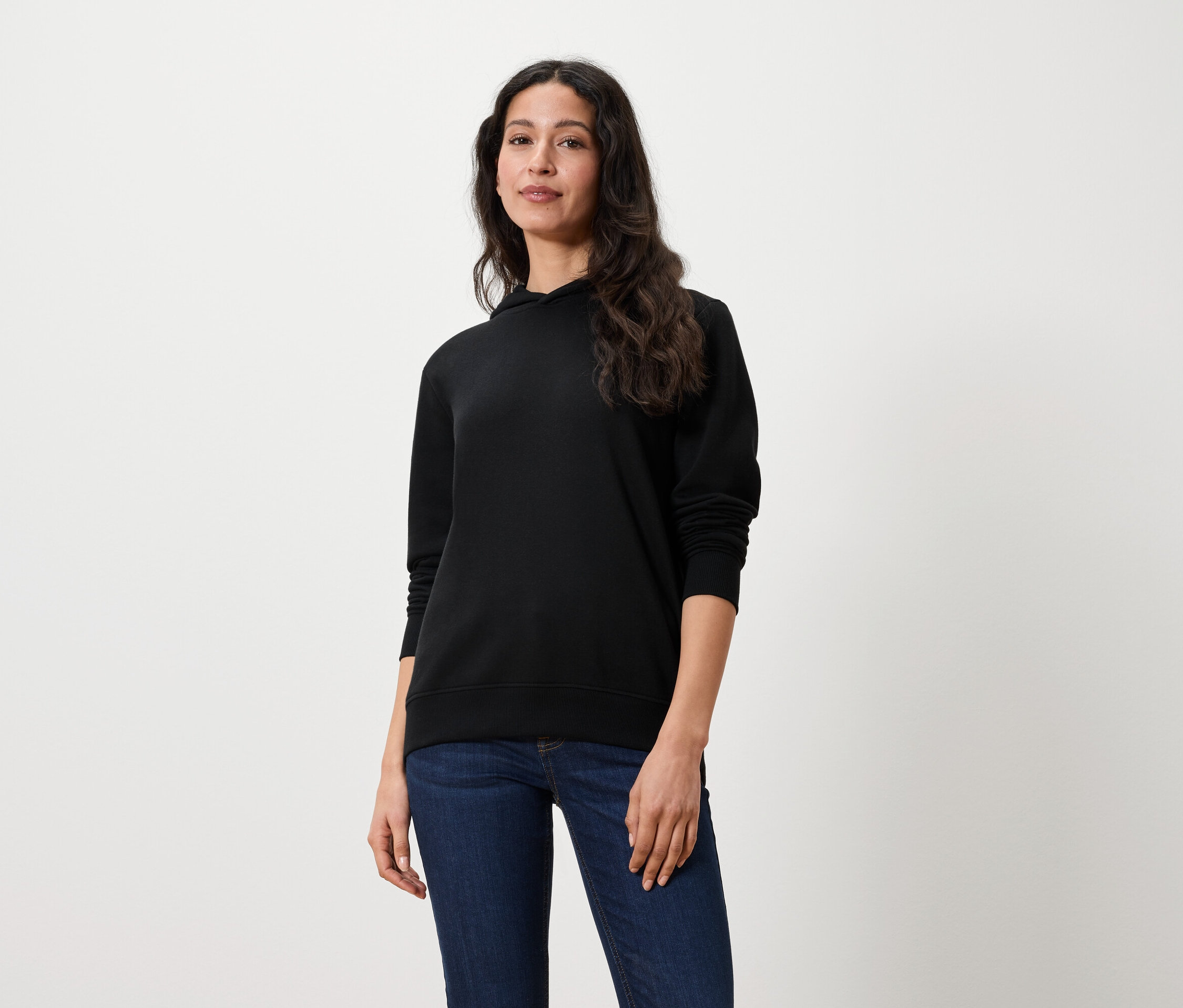 Žena s dlhými hnedými vlasmi stojí a má na sebe čiernu mikinu s kapucňou a džínsy.