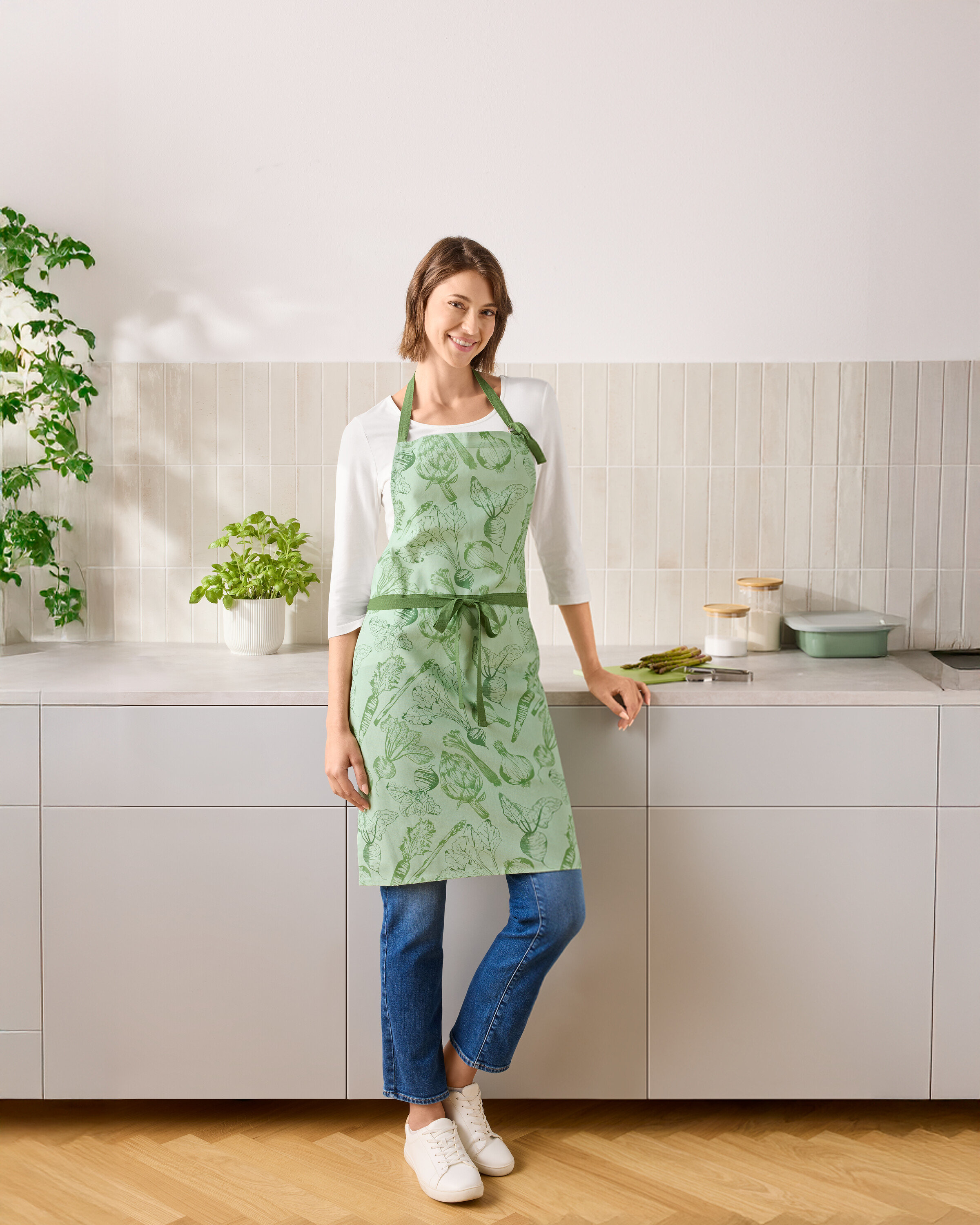 Žena stojí v kuchyni a má na sebe zelenú zásteru so zeleninou.