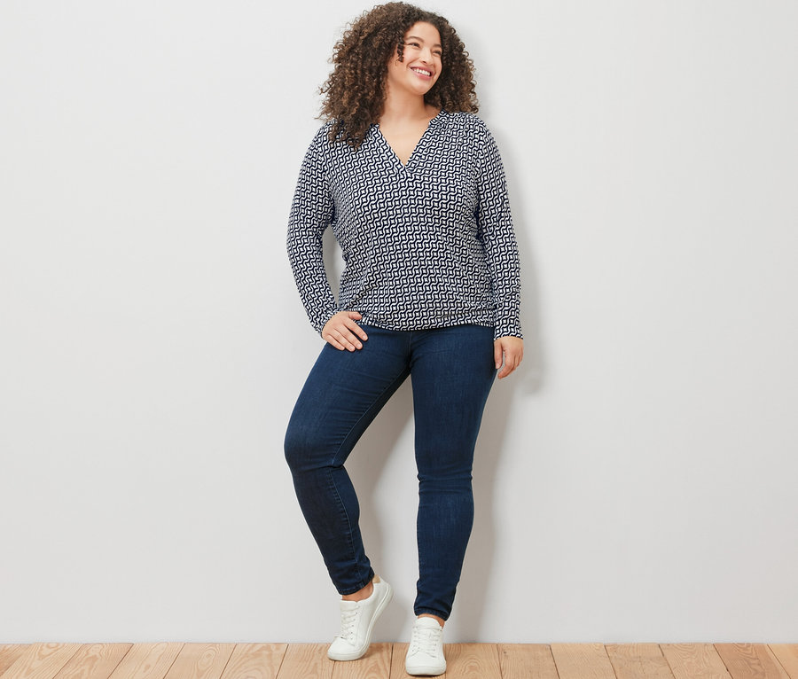 Žena stojí pred bielou stenou a má na sebe námornícke modré blúzkové tričko s potlačou, džínsy a biele tenisky.