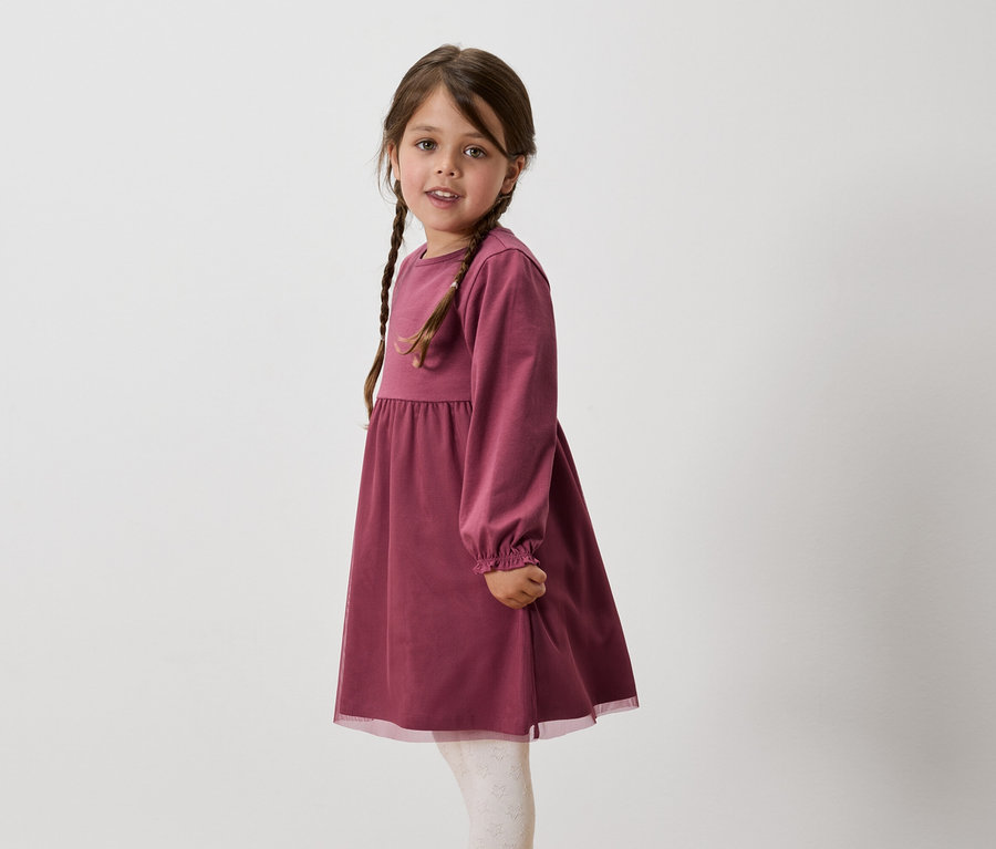 Dievča s hnedými vlasmi zapletenými do vrkočov má na sebe vínové šaty s dlhým rukávom a biele legíny.