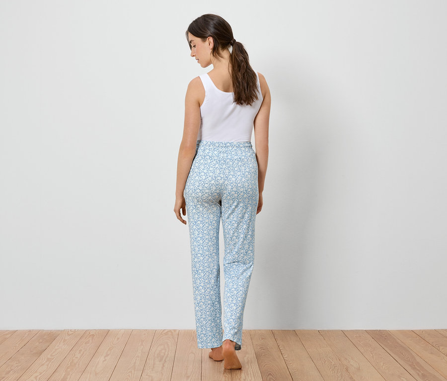 Žena zozadu, oblečená do bieleho tielka a svetlomodrých nohavíc s kvetinovým vzorom, stojí na drevenej podlahe.