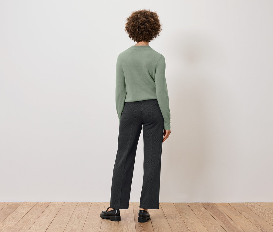 Zozadu vidíme ženu s kučeravými vlasmi v zelenom svetri a tmavých nohaviciach, stojacu na drevenej podlahe pred bielou stenou.