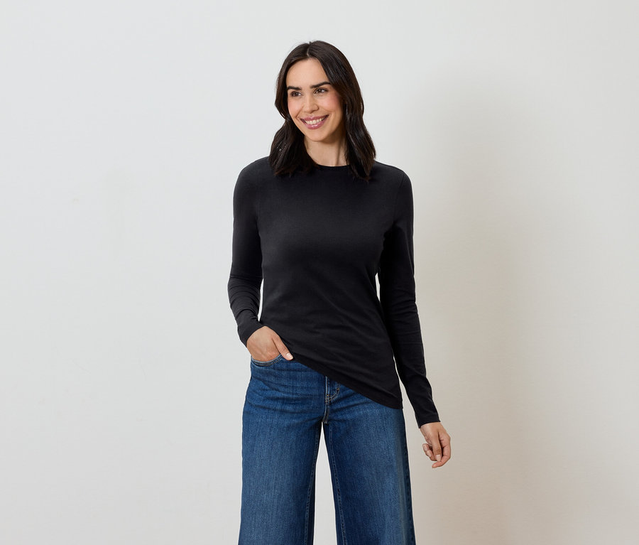 Žena s hnedými vlasmi pózuje v čiernom tričku s dlhým rukávom a modrých džínsoch.