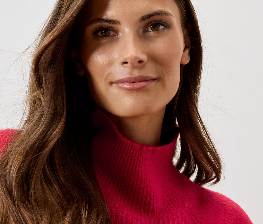 Detail tváre ženy s hnedými vlnitými vlasmi a červeným rolákom.