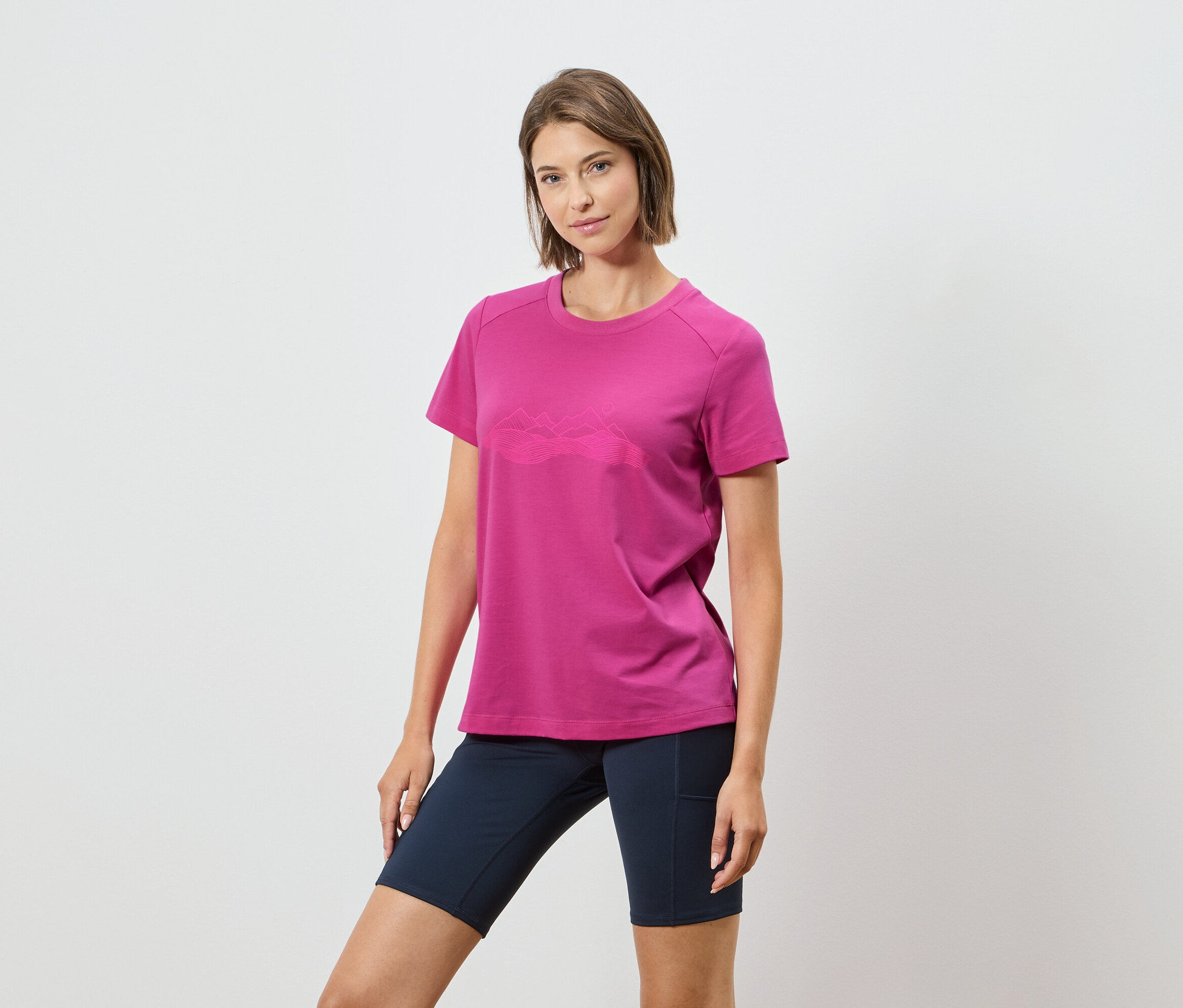 Žena má na sebe ružové tričko s horským motívom a tmavomodré šortky na bielom pozadí.