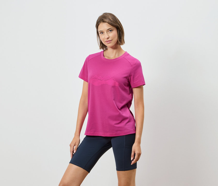 Žena má na sebe ružové tričko s horským motívom a tmavomodré šortky na bielom pozadí.