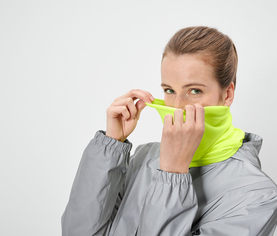 Žena si ťahá cez tvár neónovo žltý nákrčník a má na sebe sivú bundu.