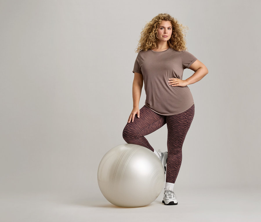 Žena s kučeravými vlasmi, v hnedom tričku a legínach so vzorom zebry pózuje s gymnastickou loptou.