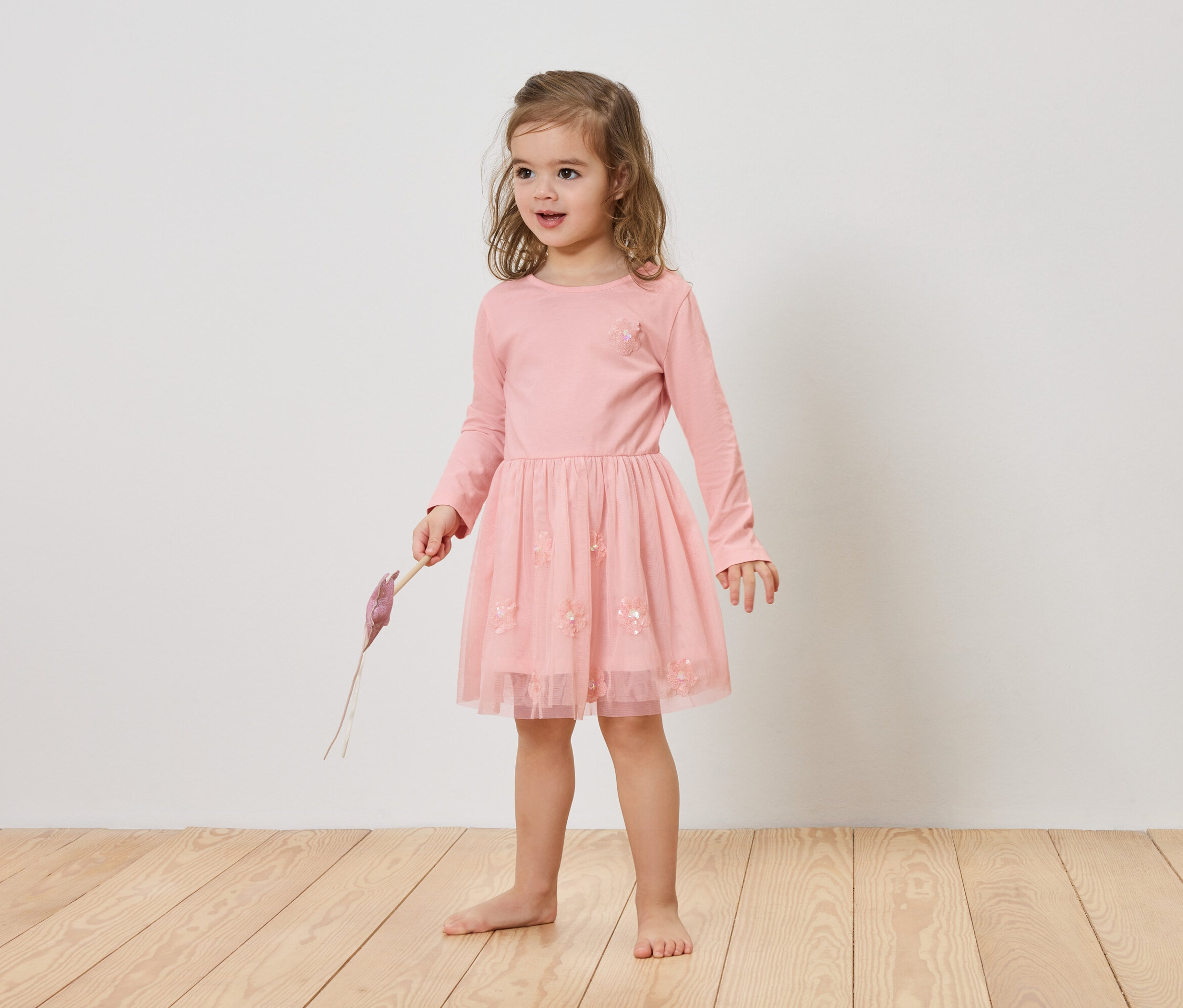 Dievča s blond vlasmi má na sebe ružové šaty a drží v ruke fialový predmet.