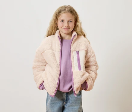 Dievča s blond kučeravými vlasmi má na sebe ružovú bundu z teddy fleece cez fialový sveter a modré džínsy.