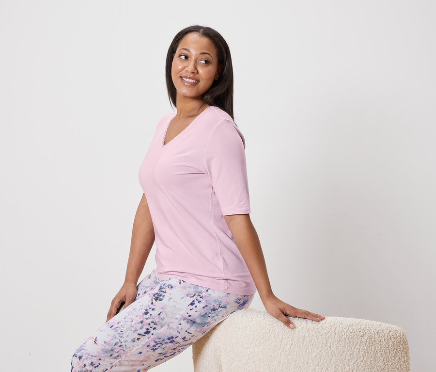 Žena sedí na stoličke a má na sebe ružový top a vzorované nohavice.