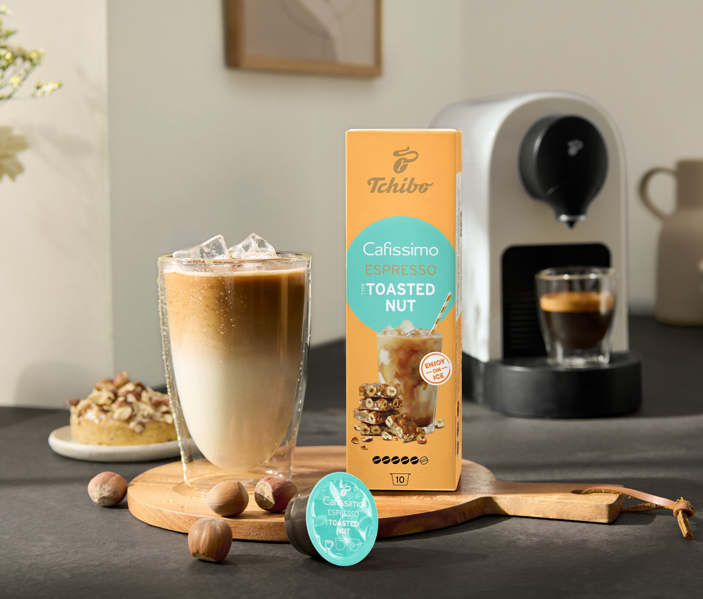 Pohár ľadovej kávy stojí vedľa balenia Cafissimo Flavoured Espresso – Toasted Nut a kávovaru Cafissimo pure+, white.