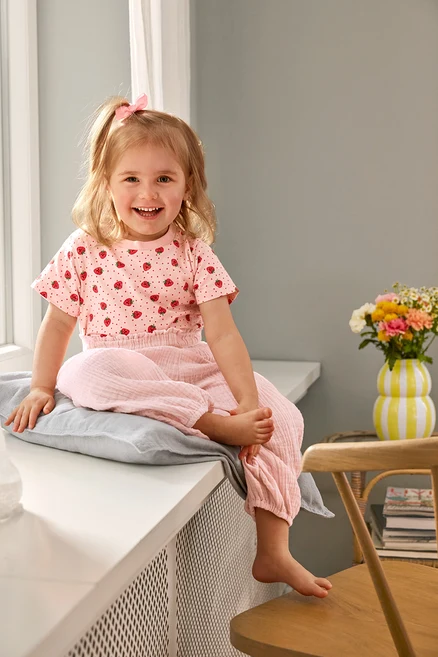 Usmievavé dievča sedí v 2 Kinder-T-Shirts a Kinder-Musselin-Hose na vankúši. Váza stojí na stoličke.