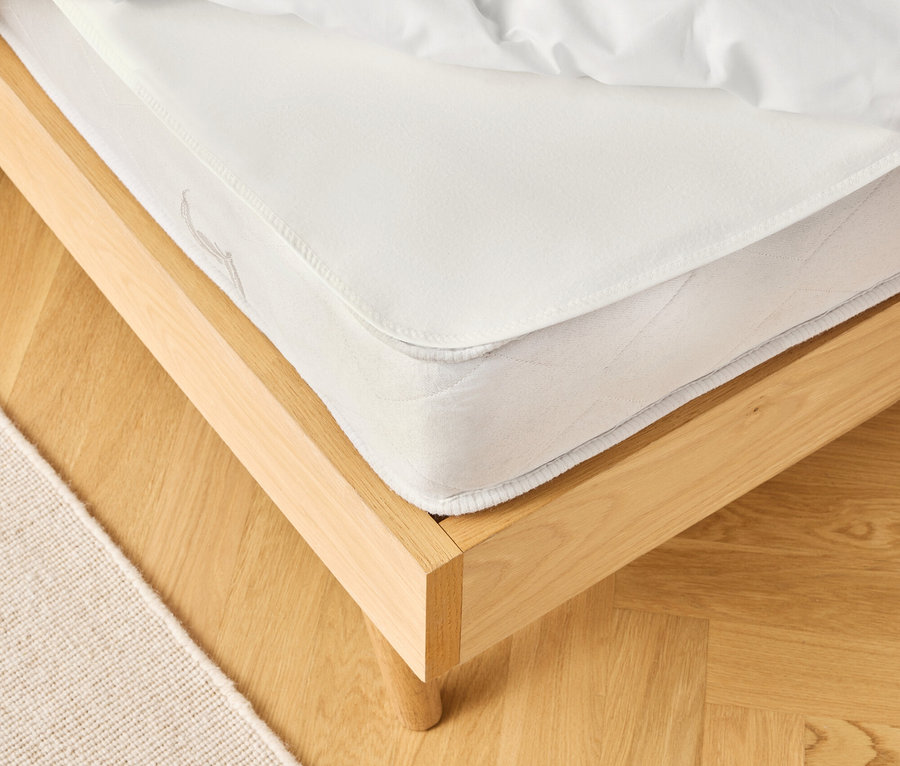 Detailný pohľad na posteľ s bielym matracom a posteľnou bielizňou na svetlej drevenej podlahe, vedľa béžový koberec.