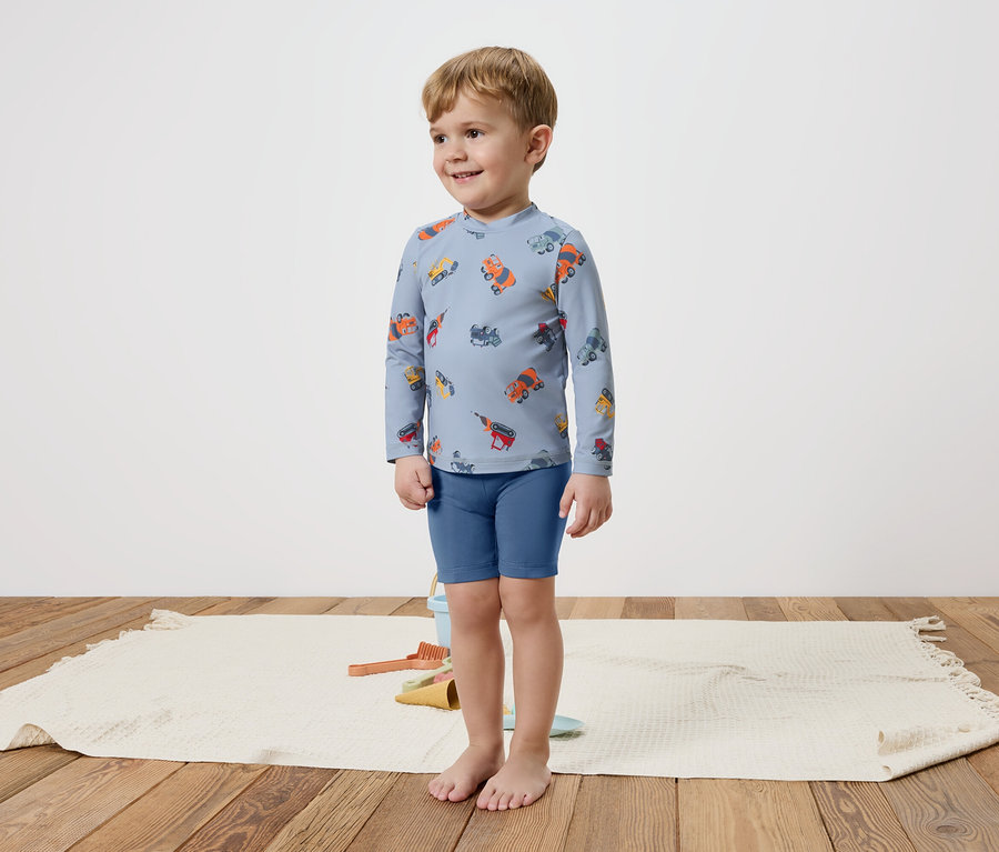 Malý chlapec stojí bosý na koberci a má na sebe modrý top s dlhým rukávom s farebnými vozidlami a modré šortky.