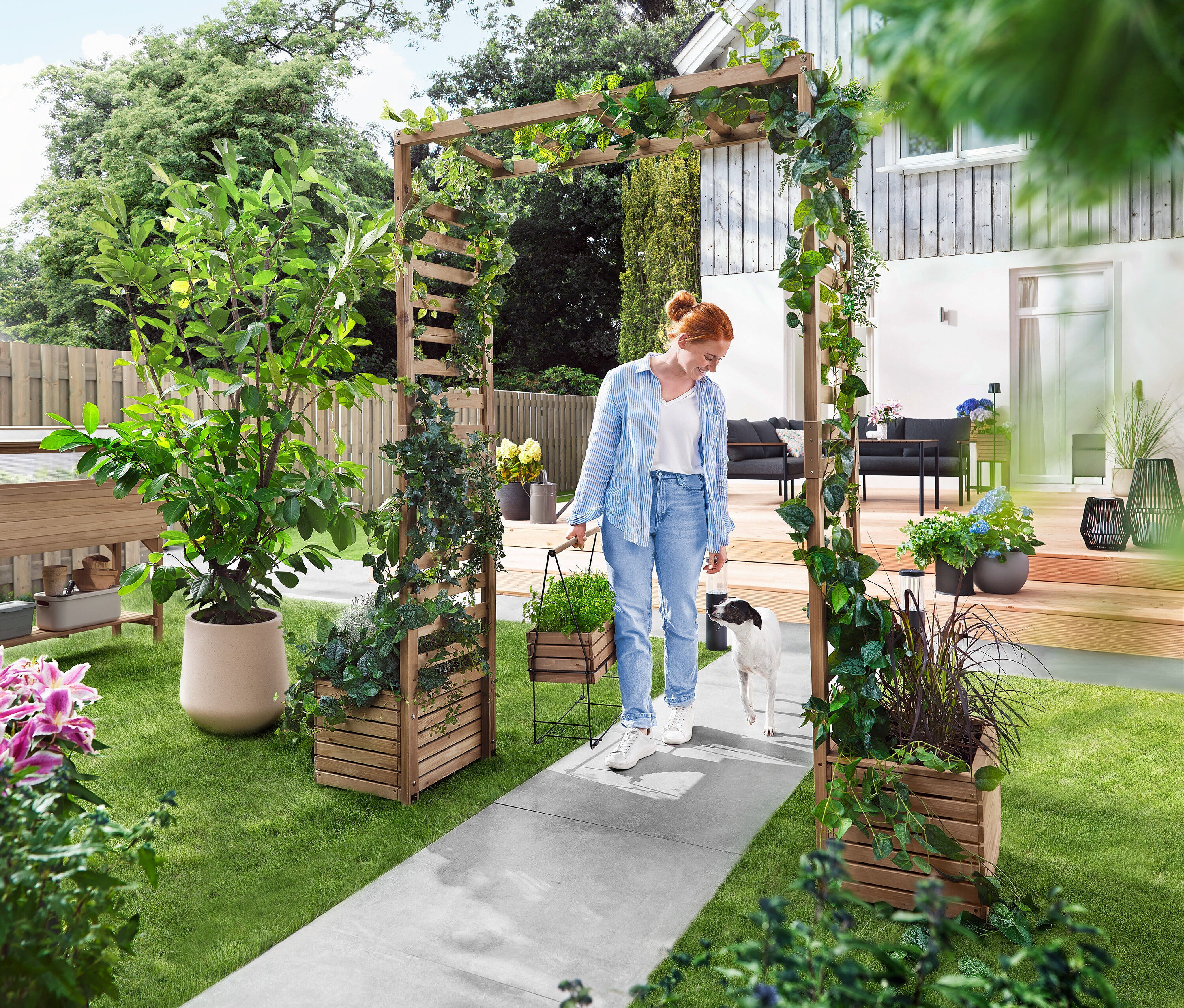 Žena prechádza so psom cez záhradný oblúk porastený rastlinami. Nesie kvetináč.