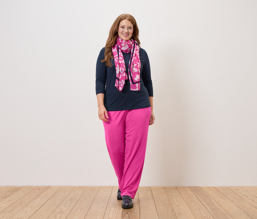 Žena s dlhými ryšavými vlasmi stojí pred bielou stenou a má na sebe tmavomodrú blúzku, ružové nohavice a ružovú šatku s kvetmi.