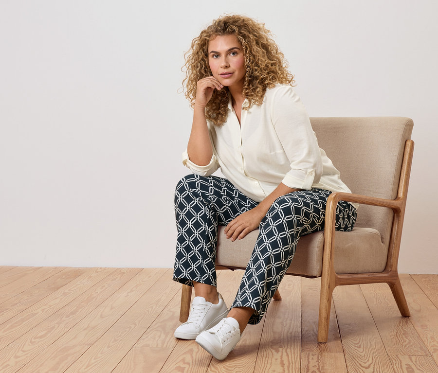 Žena s kučeravými vlasmi sedí v kresle vo vzorovaných nohaviciach a bielych teniskách.