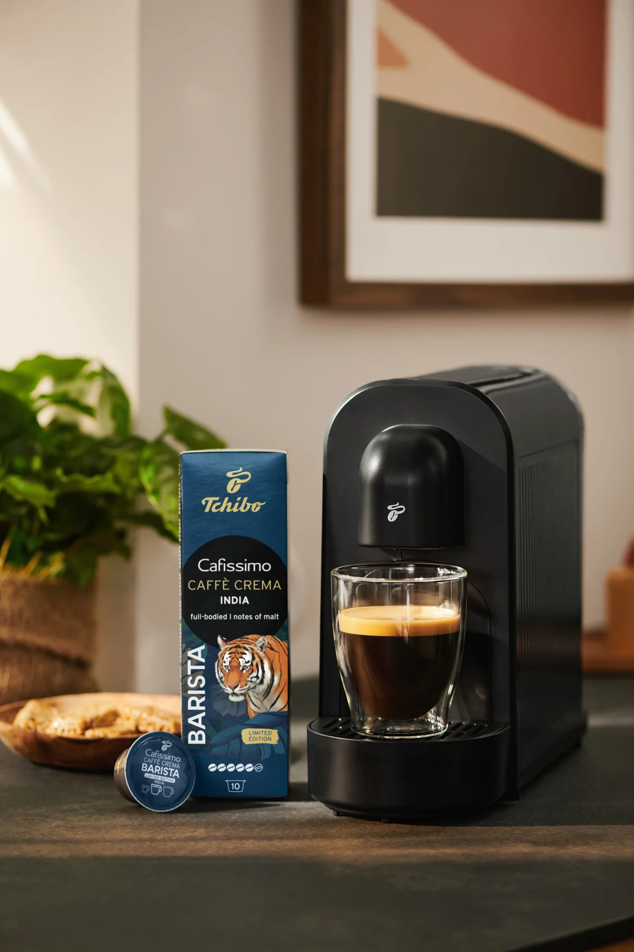 Čierny kávovar varí kávu do skleneného pohára, vedľa neho je modré balenie kapsúl Tchibo Cafissimo Caffè Crema India a jedna kapsula.