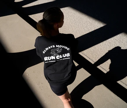 Žena zozadu v čiernej mikine s nápisom „Always Moving Run Club“ a krátkych antracitových športových legínsach »ActivePower« na ceste s tieňmi.