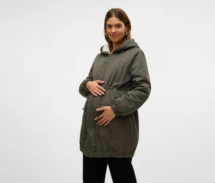 Tehotná žena v zelenej parke s kapucňou lemovanou bielou umelou kožušinou a sťahovacou šnúrkou v páse, držiaca si brucho. Má na sebe aj čierne nohavice.