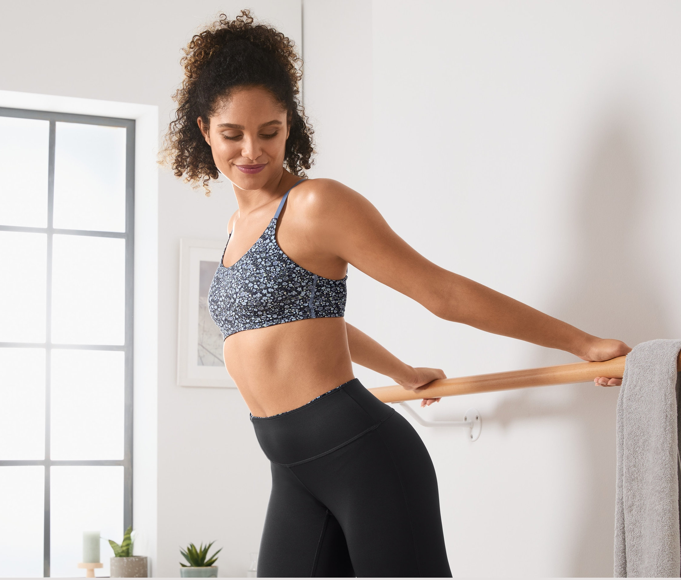 Žena sa naťahuje na baletnej tyči a má na sebe športovú podprsenku s celoplošnou potlačou.