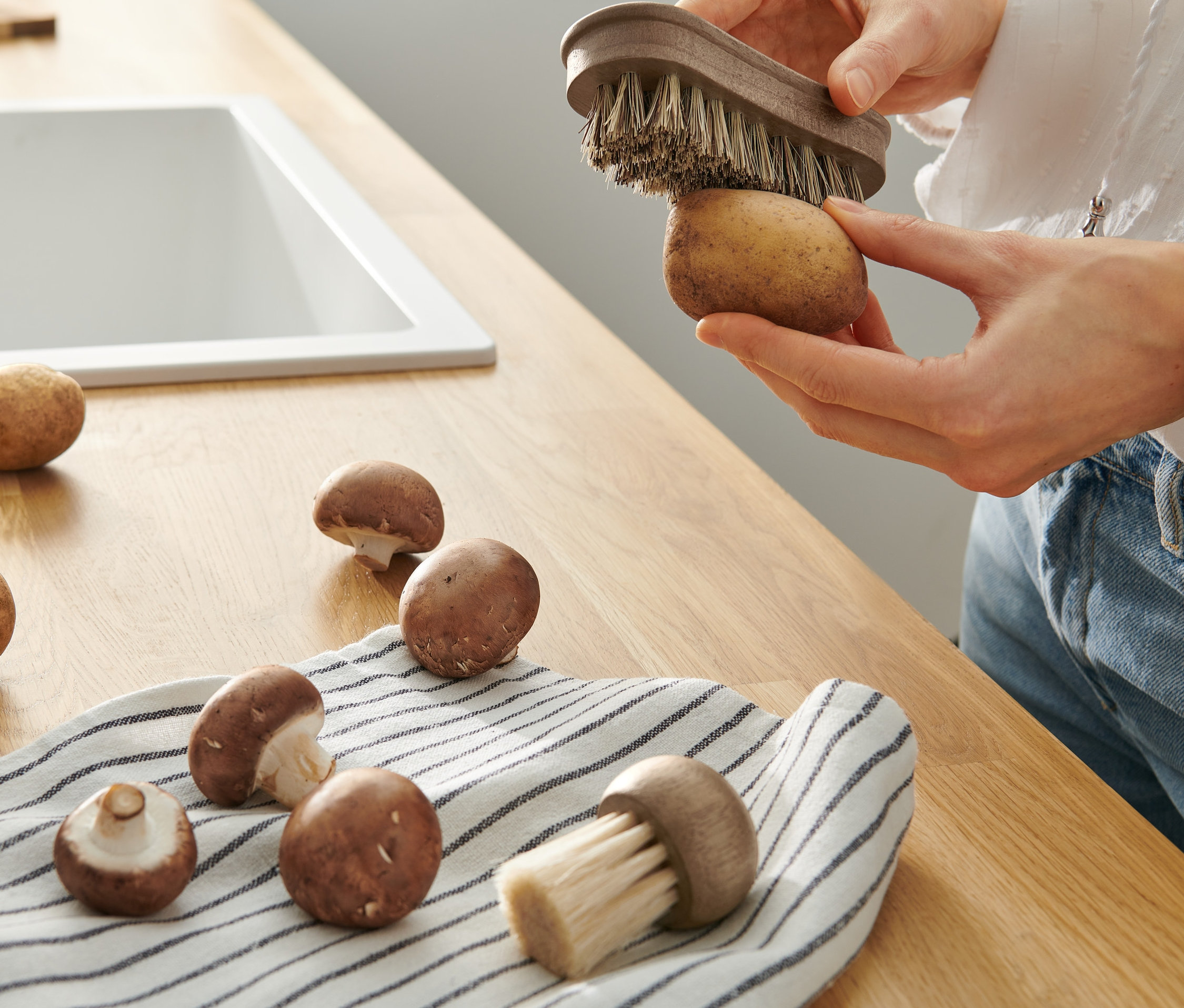 Žena čistí zemiak kefkou na zeleninu z 2 ks sady. Na linke ležia ďalšie zemiaky a huby.