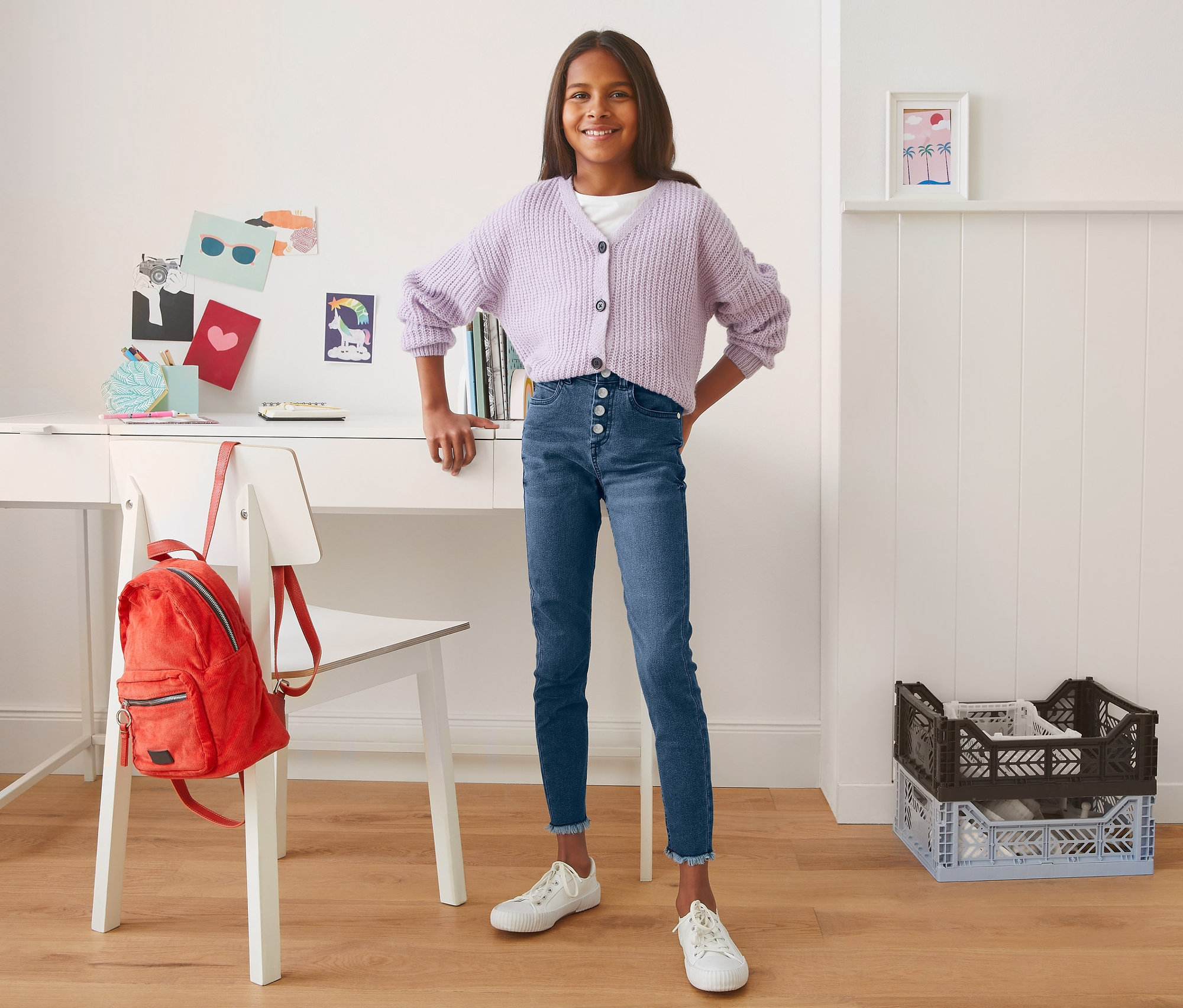 Usmievajúce sa dospievajúce dievča pózuje v modrých džínsoch s vysokým pásom a fialovom svetri pred stolom.