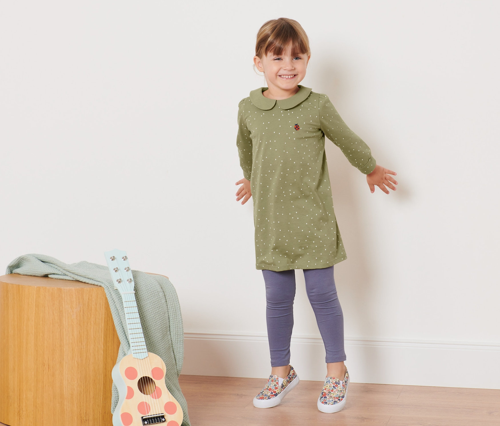 Usmievajúce sa dievčatko pózuje v olivovozelených džersejových šatách s golierom a bodkami.
