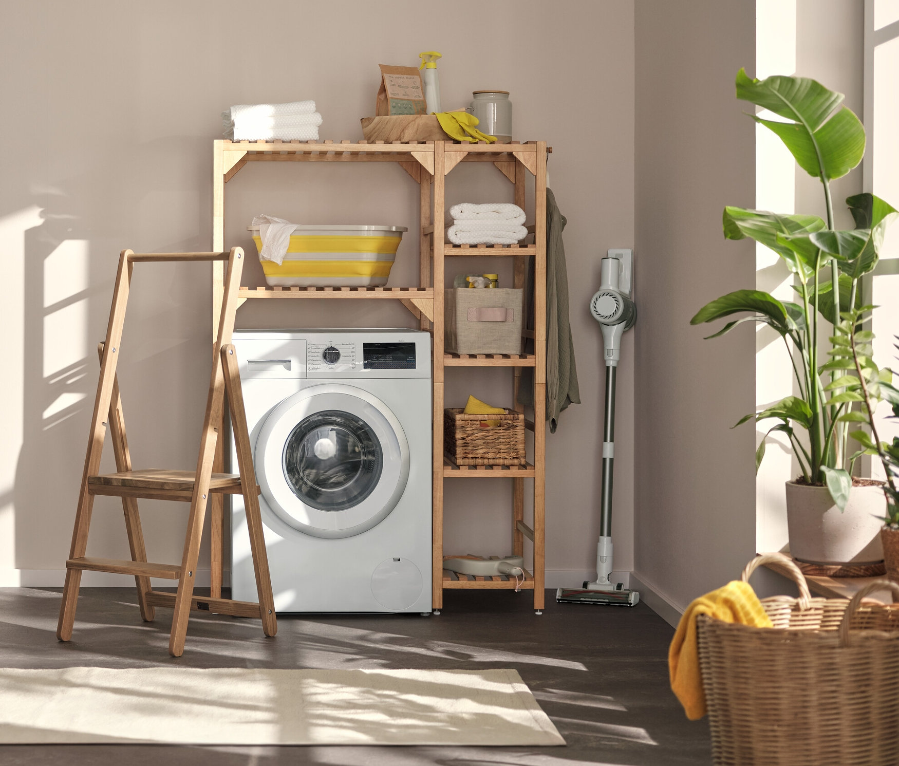 V práčovni stojí práčka, nad ňou regál s priestorom na pračku a bočným prvkom. Vľavo je nástenná polica s flexibilnými háčikmi a dnom a malý rebrík z masívneho dreva.