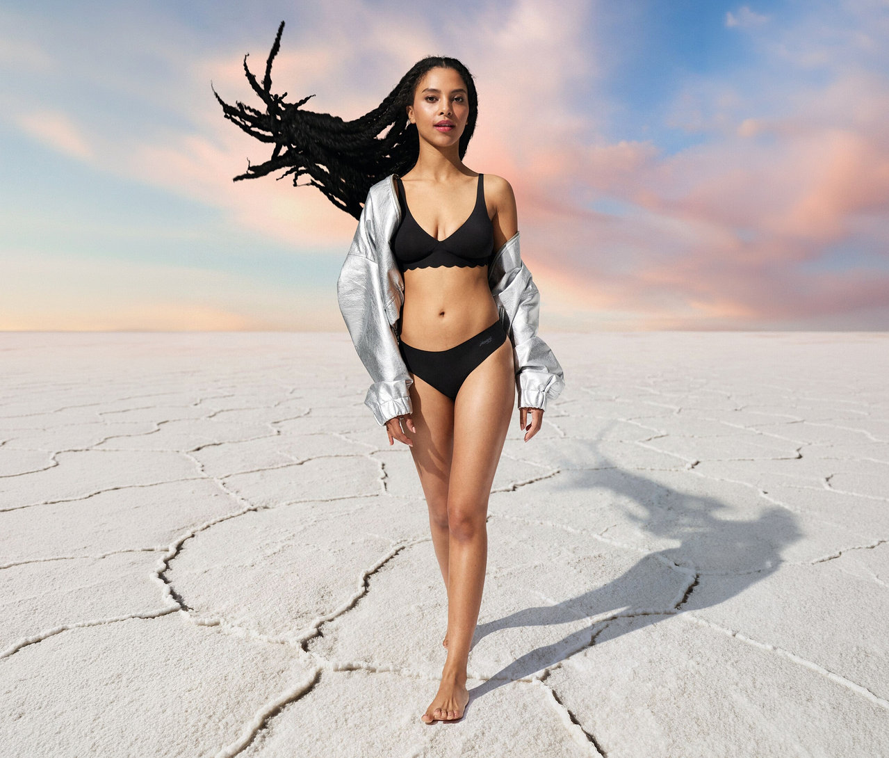 Žena v striebornej bunde a čiernej podprsenke sloggi ZERO Microfibre 2.0 Soft bra na bielom pozadí.