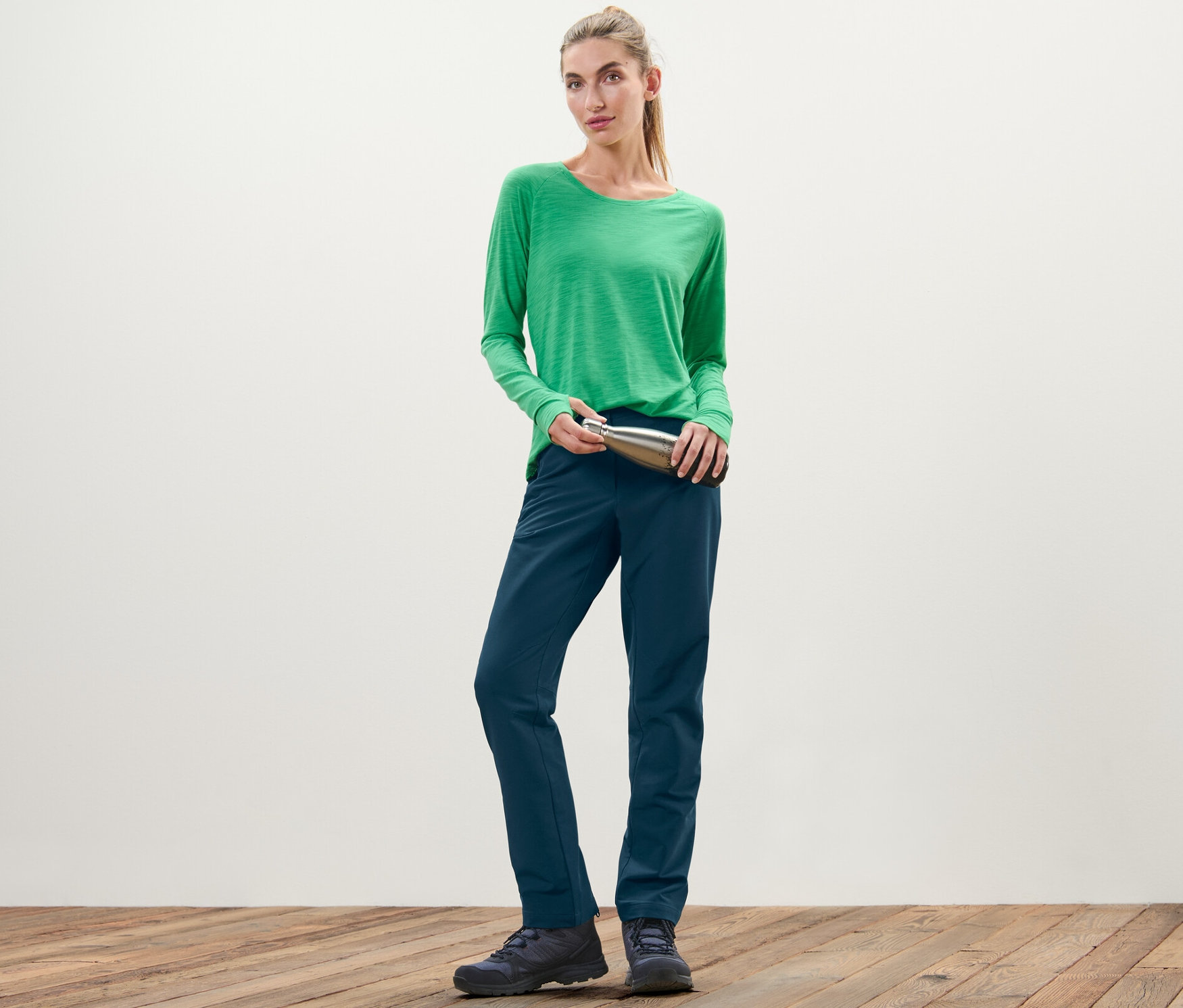 Žena v zelenom tričku drží striebornú fľašu a má na sebe softshellové nohavice.