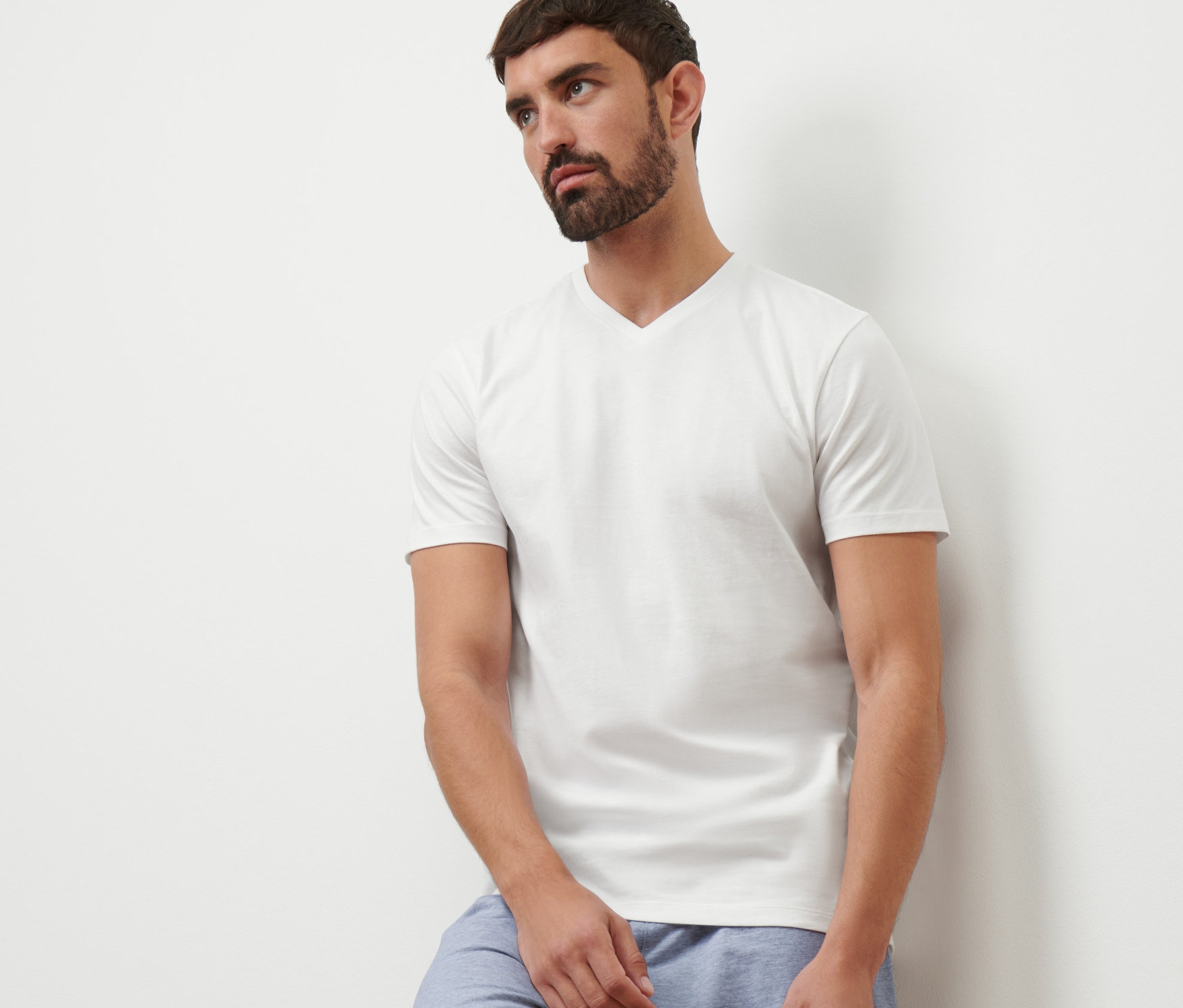 Muž opretý o stenu má na sebe biele tričko s výstrihom do V a modré šortky.