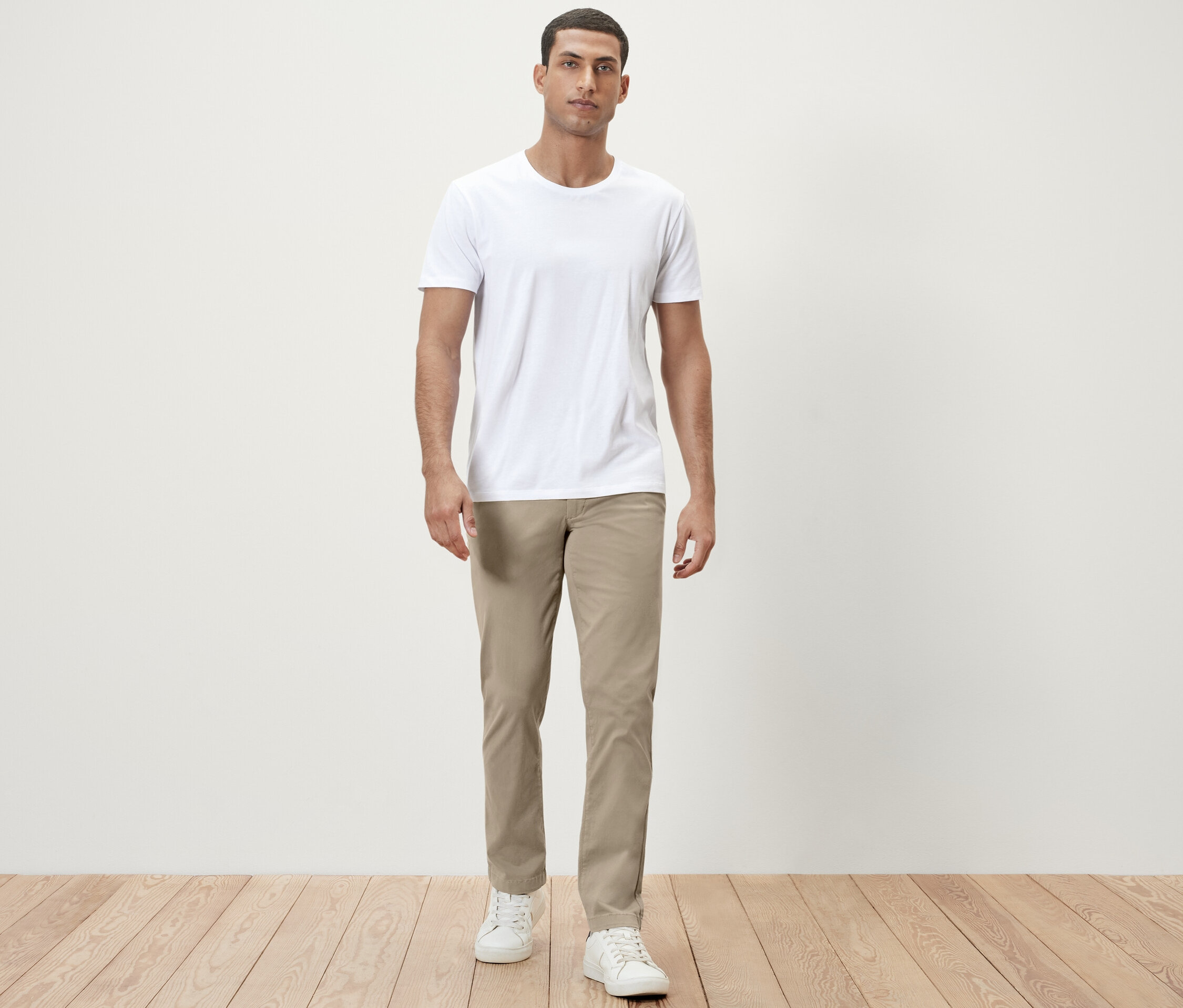 Muž stojí v bielom tričku a béžových chino nohaviciach.