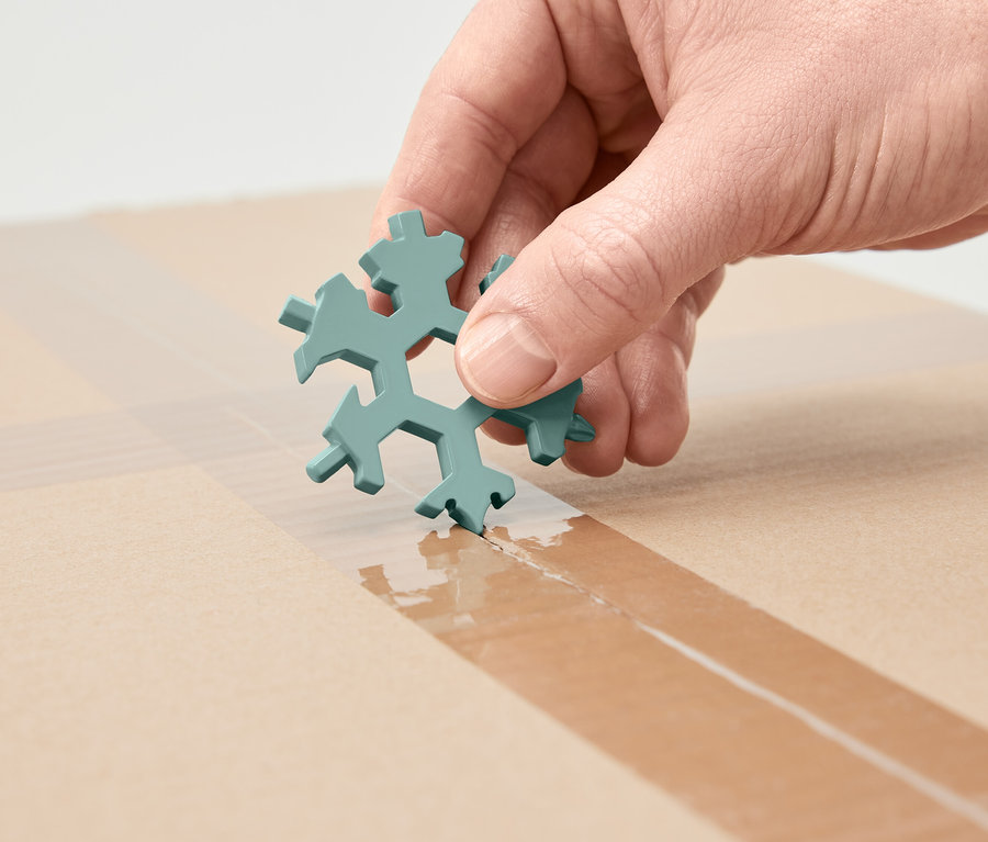 Ruka drží multifunkčný kľúč v tvare snehovej vločky a reže s ním lepiacu pásku na kartónovej krabici.