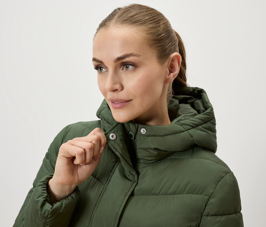Žena si zapína olivovozelenú prešívanú bundu s kapucňou.